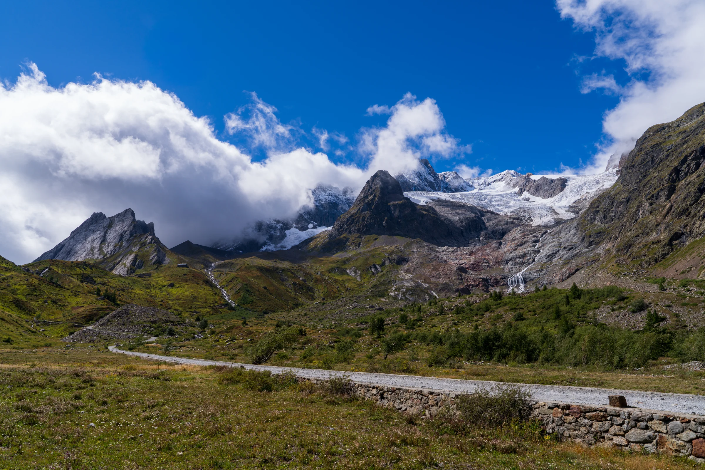 Sunlit mountain slopes and craggy peaks beneath blue sky and large white clouds.