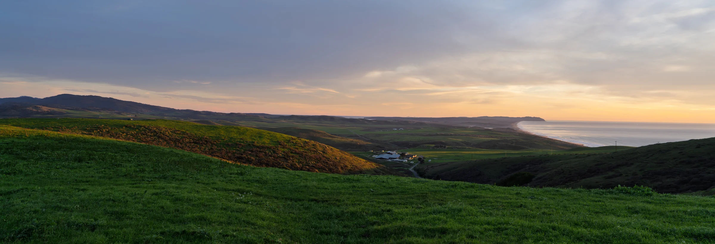 A wide view of green rolling hills beneath a gray sky with pale gold light at the horizon.