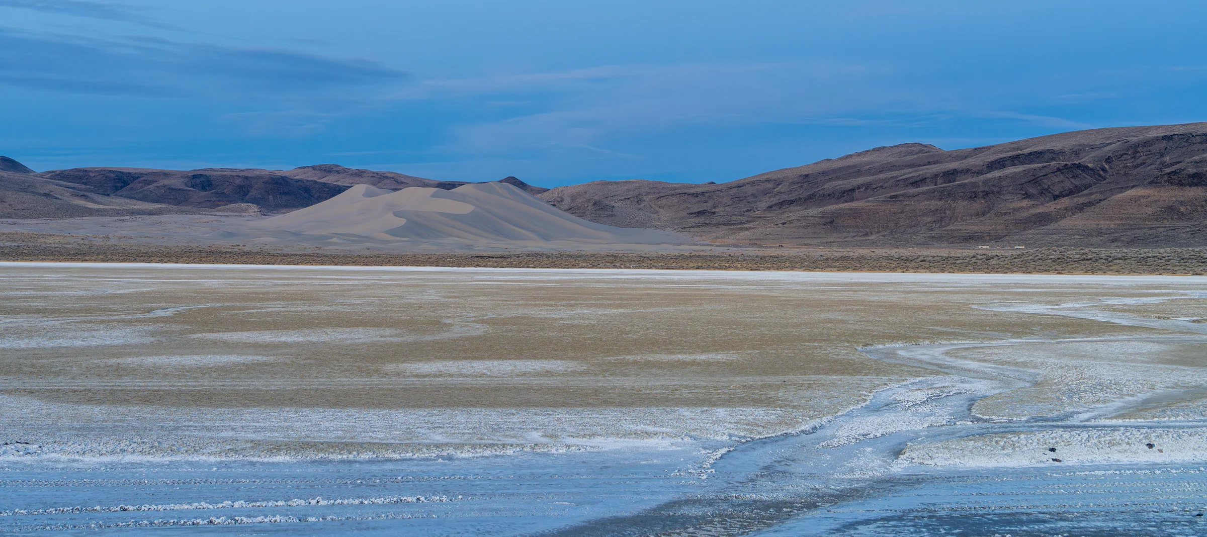 A broad pale basin crossed by shallow water with low hills and a blue-gray sky beyond.