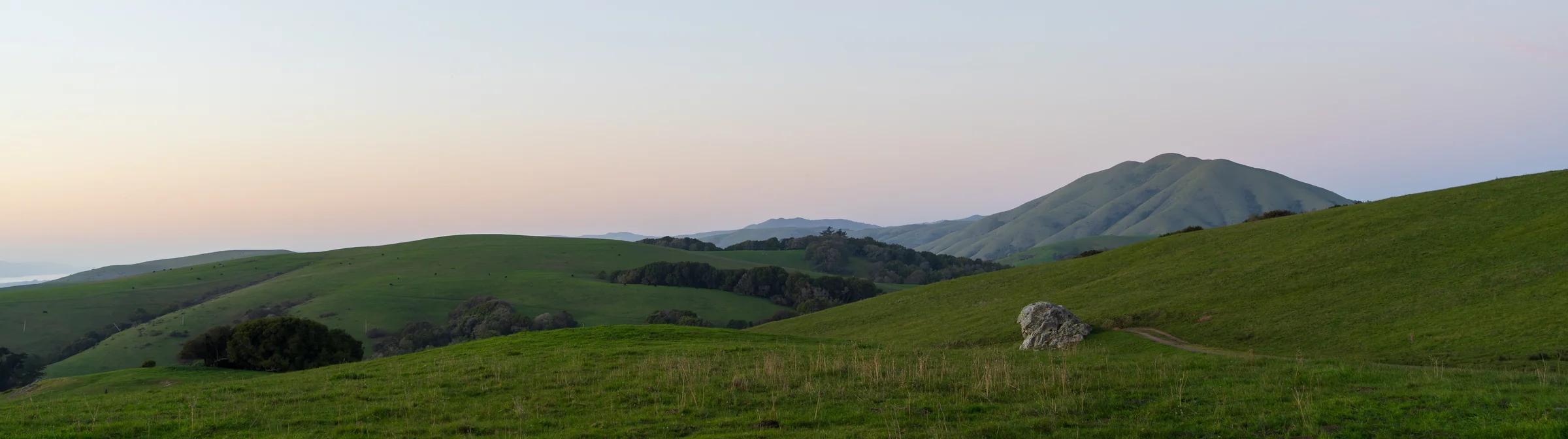 Panoramic green hills leading toward a distant ridge beneath a softly colored sky.