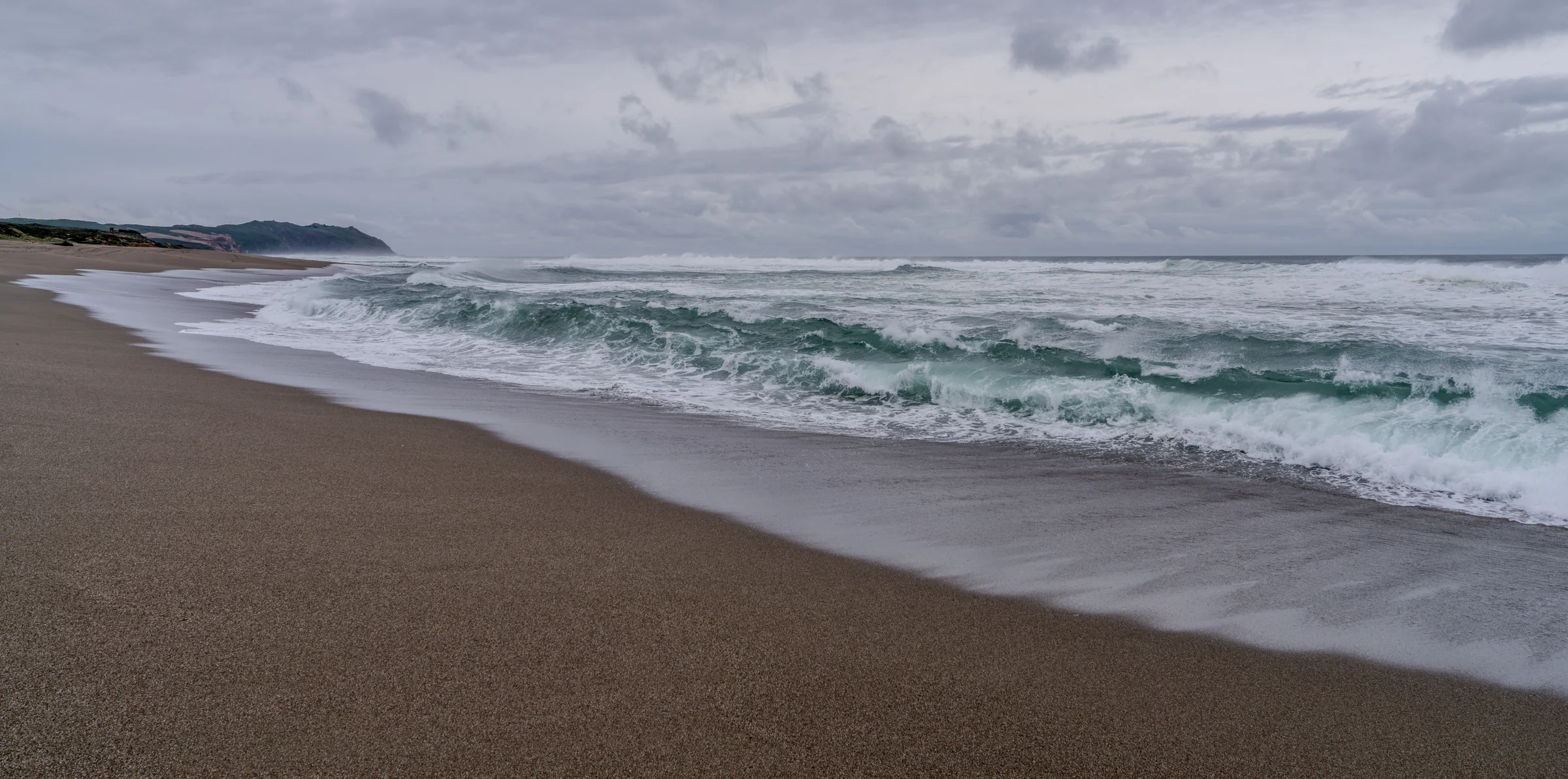 White surf rolling onto a broad sandy beach beneath heavy gray clouds.