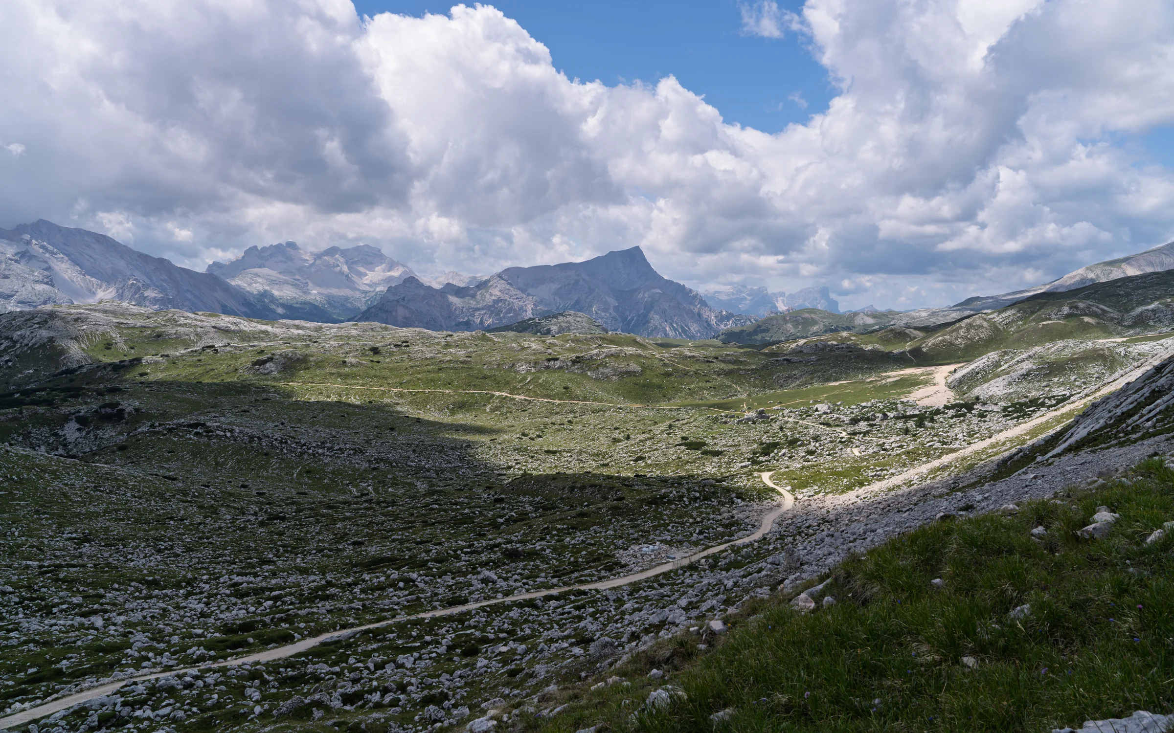 An open rocky and grassy mountain valley crossed by a winding pale trail beneath large clouds.