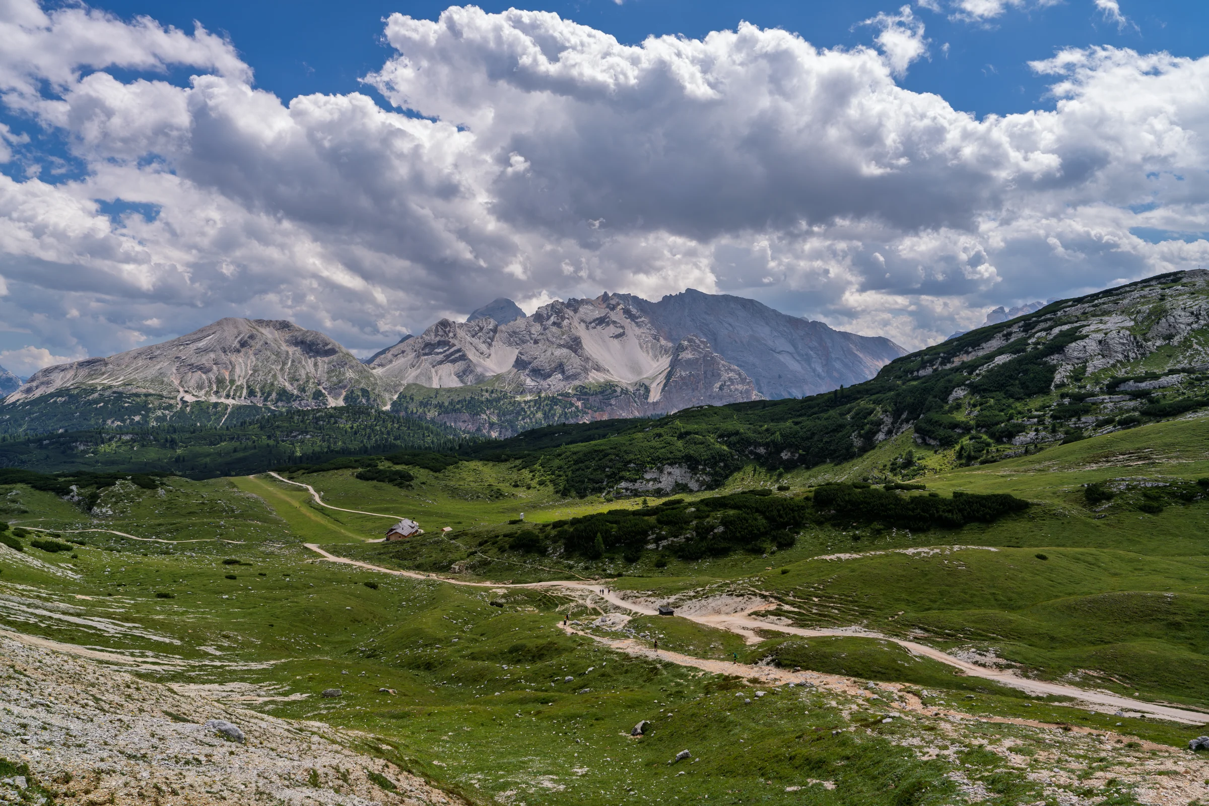 Pale paths crossing green mountain slopes with rugged peaks and white clouds in the distance.