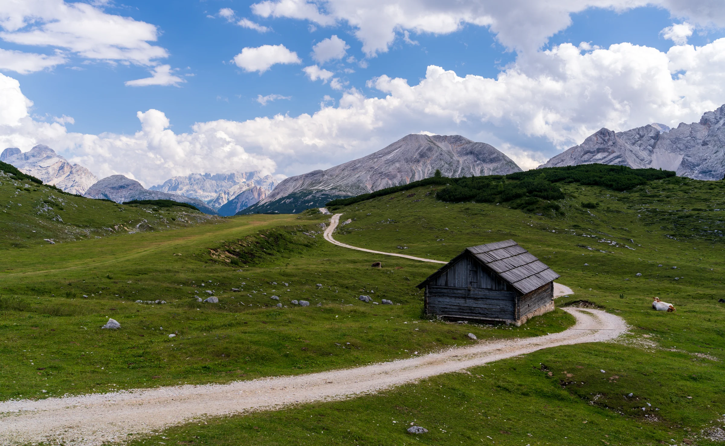A small wooden cabin beside a pale winding path, surrounded by green slopes and distant mountains.