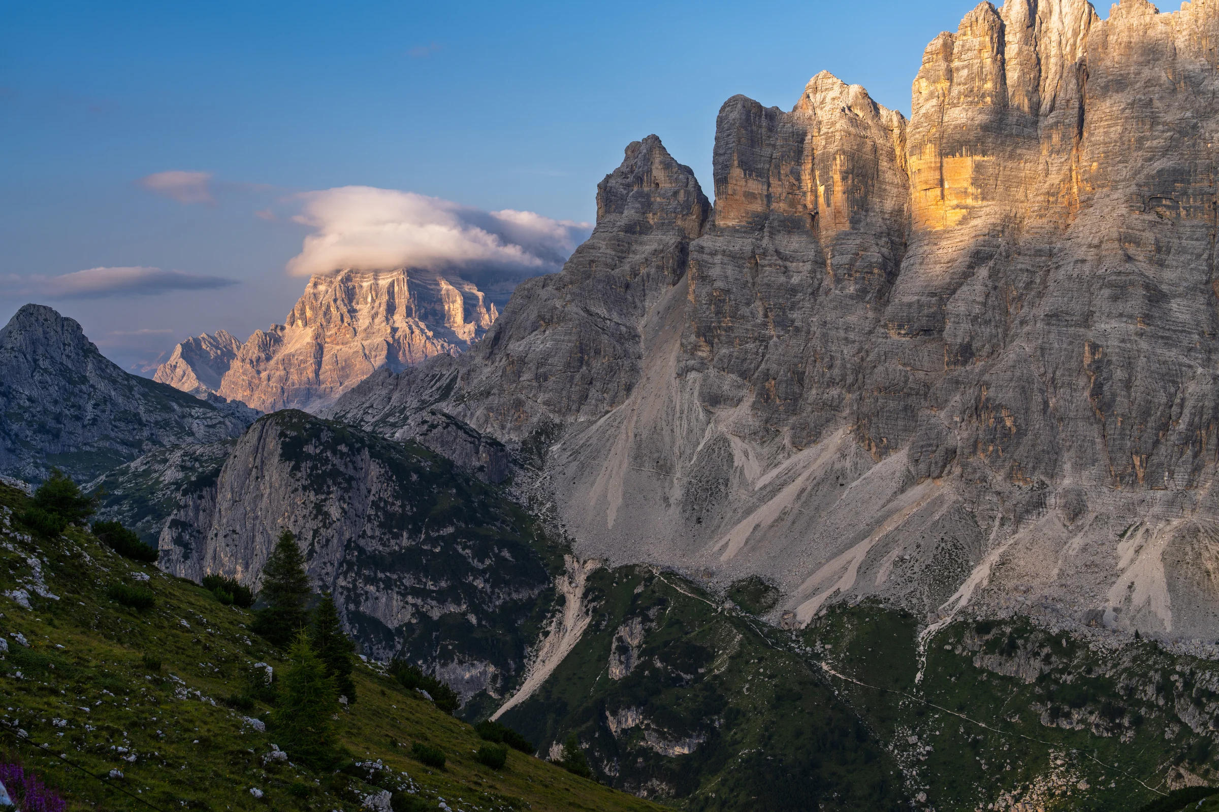 Golden light on tall mountain rock faces above a shadowed valley and a cloud-covered peak.
